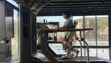 Bir kadın, robotik dış iskelet (ekzoskeleton) desteğiyle bir yürüyüş rehabilitasyon cihazında adım atarken, modern ve geniş pencereli bir ortamda fiziksel terapi çalışması yapıyor.