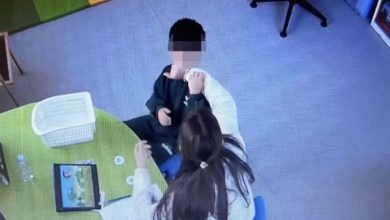 Güvenlik kamerası açısından çekilen görselde bir çocuk ve yanında oturan bir yetişkin görülüyor. İç mekânda masa başında gerçekleşen bir etkileşim anı kaydedilmiş; masanın üzerinde tablet ve çeşitli materyaller bulunuyor. Görüntü, eğitim veya bakım ortamını andıran bir alanı gösteriyor.