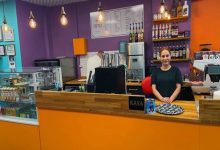 Renkli bir kafe ortamında tezgâhın arkasında duran bir kişi kameraya bakıyor. Mor ve turuncu tonların hâkim olduğu mekânda kahve ekipmanları, içecek şişeleri ve solda cam vitrin içinde ürünler yer alıyor. Genel görünüm küçük ve canlı bir kafe atmosferi oluşturuyor.