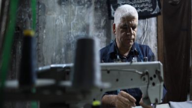 Görselde, beyaz saçlı bir kişi bir dikiş makinesinin başında çalışırken görülüyor. Loş ışıklı ve mütevazı bir atölye ortamında çekilen fotoğrafta kişi işine odaklanmış durumda. Ön planda bulanık dikiş ekipmanları yer alırken, arka plandaki yıpranmış duvarlar çalışma koşullarının zorluğunu yansıtıyor. Görüntü, emek ve geçim mücadelesini öne çıkaran bir sahne sunuyor.