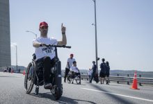 Yarış numarası göğsünde bulunan tekerlekli sandalye kullanan bir kişi önüne monte edilmiş elektrikli bir aparatla ilerlerken elini kaldırıp işaret veriyor; arka planda ise diğer katılımcılar ve görevliler görülüyor.