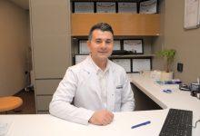 Fotoğrafta bir sağlık kuruluşundaki muayene odasında oturan bir uzman görülüyor. Beyaz doktor önlüğü ve açık renk gömlek giyen kişi, çalışma masasının arkasında kameraya dönük şekilde gülümsüyor. Arka planda çeşitli sertifika ve başarı belgelerinin yer aldığı raflar bulunuyor. Masanın üzerinde bilgisayar klavyesi, kalemler, evraklar ve küçük bir saksı bitkisi dikkat çekiyor. Ortam düzenli, aydınlık ve profesyonel bir klinik atmosferi yansıtıyor.