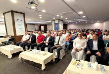 Görselde konferans veya seminer salonunda düzenlenen bir etkinlik görülüyor. Ön sırada takım elbiseli ve resmi kıyafetli katılımcılar otururken, arka sıralarda kalabalık bir dinleyici grubu yer alıyor. Salonda projeksiyon cihazı ve toplantı düzeni dikkat çekiyor; ortam eğitim ve bilgilendirme toplantısı atmosferi taşıyor.