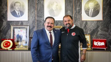 Görselde iki kişi bir makam odasında yan yana durarak kameraya gülümsüyor. Soldaki kişi takım elbise, sağdaki kişi ise üzerinde Türk bayrağı amblemi bulunan spor kıyafeti giyiyor. Arka planda duvarda çerçeveli portreler, Türk bayraklı plaketler ve çeşitli ödül objeleri yer alıyor. Görüntü, resmi bir ziyaret veya iş birliği görüşmesi sonrasında çekilmiş hatıra fotoğrafı izlenimi veriyor.