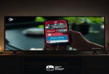 Televizyon ekranında, elde tutulan bir akıllı telefonda Türkiye Milli Paralimpik Komitesi’nin mobil uygulaması görüntüleniyor. Ekranda Paris 2024 içerikleri yer alırken, altta TMPK logosu bulunuyor.