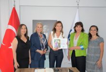 Görselde, bir kurum ofisinde gerçekleştirilen toplantı sırasında beş kadın birlikte poz veriyor. Katılımcılardan biri sertifika ve plaket benzeri bir belgeyi tutarken, arka planda Türk bayrağı ve kurum bayrağı yer alıyor. Görsel, Dünya Çevre Günü kapsamında düzenlenen asbest farkındalığı ve bilgilendirme etkinliğini yansıtıyor.