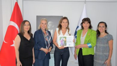 Görselde, bir kurum ofisinde gerçekleştirilen toplantı sırasında beş kadın birlikte poz veriyor. Katılımcılardan biri sertifika ve plaket benzeri bir belgeyi tutarken, arka planda Türk bayrağı ve kurum bayrağı yer alıyor. Görsel, Dünya Çevre Günü kapsamında düzenlenen asbest farkındalığı ve bilgilendirme etkinliğini yansıtıyor.
