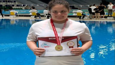 Bir yüzme havuzunun önünde çekilen fotoğrafta, boynunda altın madalya taşıyan genç bir sporcu başarı belgesini tutarak poz veriyor. Beyaz tişört giyen sporcu kameraya gülümserken, arka planda havuz ve müsabaka alanında bulunan görevliler görülüyor. Fotoğraf, yüzme yarışmasında elde edilen birincilik başarısını yansıtıyor.
