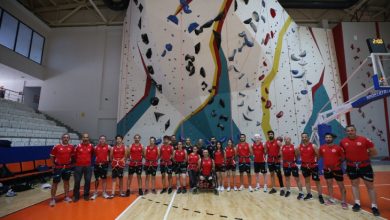 Kapalı bir spor salonundaki yüksek yapay tırmanış duvarının önünde, kırmızı formalar ve emniyet kemerleri giymiş Para Tırmanış Milli Takımı sporcuları toplu halde poz veriyor.