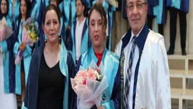Görselde, mezuniyet töreninde çekilmiş bir an yer alıyor. Ortada mezun cübbesi giymiş genç bir kadın elinde çiçek buketiyle dururken, yanında annesi ve akademik cübbe giymiş bir öğretim üyesi poz veriyor. Arka planda ise törene katılan diğer mezunlar görülüyor.