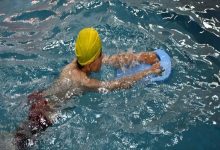 Görselde, sarı bone takan bir çocuk kapalı yüzme havuzunda mavi bir yüzme tahtasıyla su üzerinde ilerliyor. Çocuk yüzme egzersizi yaparken havuzun suyu çevresinde dalgalanıyor.