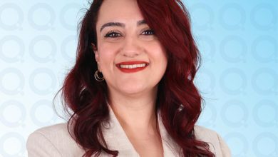 Koyu kızıl dalgalı saçlı, açık renk blazer giyen, altın halka küpeli; mavi tonlu modern bir arka planda kameraya sakin ve profesyonel bir ifadeyle bakan bir portre.