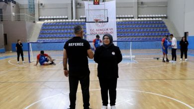 Görselde, kapalı spor salonunda bir golbol müsabakası öncesi veya sırasında hakem ile bir kadın orta sahada ayakta duruyor. Arka planda golbol oyuncuları, kale ve antrenman hazırlıkları görülürken, tribünler ve "T.C. Gençlik ve Spor Bakanlığı" yazılı pankart spor salonu ortamını yansıtıyor.