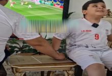 Görselde, İran milli takım forması giyen bir çocuk bir masanın kenarında otururken, yanında bulunan kişi kartondan yapılmış küçük bir futbol sahası maketi üzerinde elini hareket ettiriyor. Arka planda televizyonda futbol maçı yayınlanıyor. Sahne, çocuğun maçı dokunarak takip etmesine yardımcı olunan duygusal bir anı yansıtıyor.