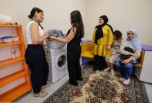 Görselde, engelli bireylerin günlük yaşam becerilerini geliştirmeye yönelik bir eğitim ortamı görülüyor. Ön planda iki kişi çamaşır makinesi başında uygulamalı çalışma yaparken, diğer katılımcılar oturma alanında etkinliği izliyor. Sınıf ortamında raflar, oturma grubu ve günlük yaşam eğitimine uygun ekipmanlar yer alıyor.