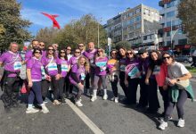 Mor tişörtler giyen kalabalık bir grup, güneşli bir günde şehir caddesinde düzenlenen bir koşu etkinliği öncesinde gülümseyerek toplu hatıra fotoğrafı çektiriyor.