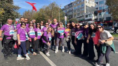 Mor tişörtler giyen kalabalık bir grup, güneşli bir günde şehir caddesinde düzenlenen bir koşu etkinliği öncesinde gülümseyerek toplu hatıra fotoğrafı çektiriyor.