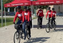 Görselde, kırmızı-beyaz formalar ve kasklar giyen tandem bisiklet sürücüleri, kırmızı çadırların önünden toplu halde hareket ediyor. Önde görme engelli sporcular ve onlara eşlik eden pilot sürücüler yer alırken, etkinlik açık havada gerçekleştiriliyor.