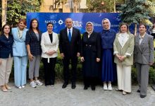 Taş döşeli açık alanda, NATO ve SETA logolarının yer aldığı büyük mavi bir afişin önünde dokuz kişilik bir grup fotoğrafı çekilmiş. Grubun ortasında, koyu renk takım elbisesiyle eski Dışişleri Bakanı Mevlüt Çavuşoğlu yer alıyor. Sağdan ikinci sırada açık renk kıyafetiyle SERÇEV Genel Sekreteri Edibe Sinem Ersoy bulunuyor. Her iki isim de kameraya dönük, resmi bir etkinlik atmosferi içinde poz veriyor. Arka planda yeşillikler ve ağaçlar görülüyor.