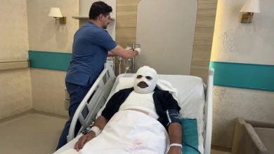 Görselde, hastane odasında tedavi gören bir hasta hastane yatağında uzanıyor. Hastanın başı ve vücudunun büyük bir bölümü bandajlarla sarılıyken, yanında bir sağlık çalışanı serum ekipmanıyla ilgileniyor. Oda, tıbbi ekipmanlar ve hasta yatağıyla donatılmış bir hastane ortamını yansıtıyor.