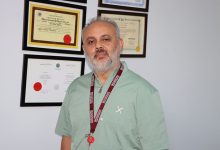 Prof. Dr. Ünal Aydın, iç mekânda, duvarda asılı çok sayıda diploma ve sertifikanın önünde duruyor. Üzerinde açık yeşil ameliyat önlüğü (scrubs) bulunuyor. Boynunda bordo renkte, üzerinde “Bezmialem Vakıf Üniversitesi” yazan bir yaka kartı askısı var. Arka plandaki çerçeveler; Sağlık Bakanlığı onaylı belgeler ve profesyonel sertifikalar gibi görünüyor. Aydın, duruşu ve ortamın düzeniyle klinik bir çalışma alanında, deneyimli bir cerrah izlenimi veriyor.