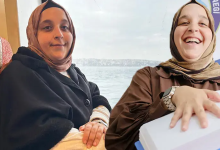 Görselde başörtülü iki kadın yan yana oturuyor. Soldaki kadın kameraya bakarken, sağdaki kadın gülümseyerek elini önündeki beyaz kutunun üzerine koyuyor. Arka planda deniz manzarası görülüyor.