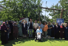Fotoğrafta, açık havada düzenlenen etkinlikte engelli anneleri ve katılımcılar toplu halde objektife poz veriyor. Ön sırada tekerlekli sandalye kullanan bir kişi yer alırken, arka planda ağaçlar, yeşil alan ve etkinliğe ait afişler görülüyor.