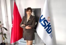 Resimde, koyu renk bir blazer ve etek takımı giyen Ayla Alkan, kolları bağlı şekilde kendinden emin bir duruşla ayakta görülüyor. Arka planda Mustafa Kemal Atatürk’ün siyah-beyaz portresi yer alıyor. Sağında USKD bayrağı, solunda ise Türk bayrağı bulunuyor. Ortam, kurumsal bir ofis veya dernek çalışma alanı izlenimi veriyor.