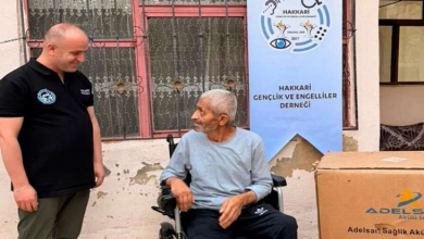 Görselde akülü tekerlekli sandalyede oturan bir kişi ile yanında duran bir görevli görülüyor. Arka planda Hakkari Gençlik ve Engelliler Derneği afişi, sağ tarafta ise tekerlekli sandalyeye ait büyük bir kutu yer alıyor.