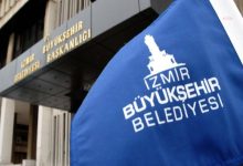 Bir binanın önünde mavi renkli "İzmir Büyükşehir Belediyesi" yazan bayrak var. Binanın üstündeki yazı tam olarak seçilmiyor.