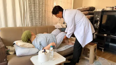 Görselde sağlık görevlisi, ev ortamında koltukta uzanan bir kişiye bacak bölgesinden muayene ve bakım uyguluyor.
