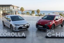 Sahile yakın açık alanda yapılan bir tanıtım alanında Toyota marka iki araç yan yana sergileniyor. Solda gümüş renkli 2026 Corolla, sağda kırmızı 2026 C‑HR bulunuyor. Her iki modelin önünde şeffaf tanıtım panoları yer alıyor ve arka planda palmiye ağaçları ile modern bir Toyota binası görünüyor.