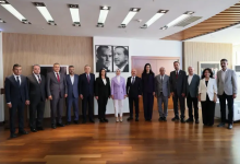 Modern bir toplantı salonunda yan yana dizilmiş kalabalık bir protokol grubu görülüyor. Ortada Aile Bakanı, yanında Aydın Valisi ve Aydın Belediye Başkanı olmak üzere toplamda on üç kişi resmi bir poz veriyor. Her biri takım elbise ve profesyonel kıyafetlerle, arka planda ise duvarda asılı iki adet Atatürk portresi bulunuyor. Ortam resmi, düzenli ve protokol buluşması niteliğinde.