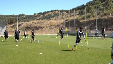 Görselin üst bölümünde, açık ve güneşli bir havada dağlık ve ağaçlık bir tepenin eteğindeki çim sahada antrenman yapan Ampute Futbol Milli Takımı oyuncuları yer alıyor. Ön planda, siyah antrenman takımı giymiş ve kanedyen (koltuk değneği) kullanan bir futbolcu, sahaya dikilmiş dikey sarı slalom çubuklarının arasından geçerek çeviklik çalışması yapıyor. Sahaya yayılmış diğer millî futbolcular ise küçük beyaz antrenman engelleri ve huniler eşliğinde farklı istasyonlarda ısınma ve koordinasyon hareketlerini sürdürüyor; arka planda yüksek tel örgüler ve tepe yamacı görülüyor.