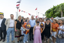 Görselde Mersin Büyükşehir Belediye Başkanı, engelli gençler ve aileleriyle birlikte açık alanda toplu fotoğraf çekiminde görülüyor. Başkan, grubun ortasında yer alıyor; etrafında turuncu tişörtleriyle “Engelliler Spor Kulübü” üyeleri ve aileleri bulunuyor. Katılımcıların bir kısmı kulübün logosunun yer aldığı yuvarlak pankartları taşıyor. Arka planda Türk bayrakları, ağaçlar ve etkinlik çadırları yer alıyor. Atmosfer sıcak, destekleyici ve toplumsal katılım vurgusuyla bir etkinlik ortamını yansıtıyor.
