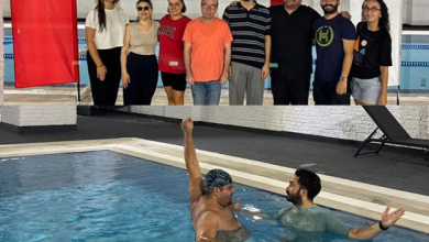 Alt alta yer alan 1. üstteki görsel: Kapalı bir havuz alanında, sekiz kişilik bir grup yan yana duruyor. Arkalarında kırmızı-beyaz dikey panolar ve havuz kenarı görülüyor. Grup, etkinlik veya eğitim ortamında bir araya gelmiş izlenimi veriyor. 2. alttaki görsel: Havuz içinde iki kişi bulunuyor. Bone takan kişi kolunu yukarı kaldırmış; yanındaki kişi ona destek veriyor veya yüzme eğitimi veriyor gibi görünüyor. Sahne, yüzme dersi ya da su terapisi uygulamasını yansıtıyor.