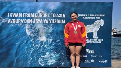 Görselde, kırmızı Galatasaray eşofman üstü ve siyah şort giyen, boynunda madalya bulunan Selin Zülal Önal, Çanakkale Boğazı’nı yüzerek geçmesinin ardından “I SWAM FROM EUROPE TO ASIA” yazılı etkinlik panosunun önünde gururla poz veriyor.