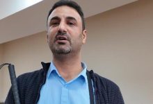 Görme engelli Dr. Önder İşlek, kapalı bir mekânda ayakta duran, kısa saçlı ve hafif sakallı bir yetişkin erkek olarak görülüyor. Açık mavi gömlek ve koyu renk dokulu bir ceket giyiyor. Sağ elinde baston tutuyor; duruşu sakin ve kendinden emin. Arka planda sade, bej tonlarında bir duvar bulunuyor.