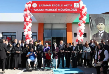 Görselin üst bölümünde, yeni bir merkezin açılış töreninde kurdele kesmek üzere yan yana dizilmiş protokol üyeleri, davetliler ve engelli bireyler yer alıyor. Giriş kapısının üzerinde kırmızı tabela zemininde beyaz harflerle "T.C. BATMAN ENGELLİ AKTİF YAŞAM MERKEZİ MÜDÜRLÜĞÜ" yazısı ve bakanlık logoları bulunuyor; kapının iki yanı kırmızı ve beyaz balonlardan yapılmış kemer sütunlarla süslenmiş durumda. Binanın sağ dış cephesinde ise kalpaklı dev bir Mustafa Kemal Atatürk posteri asılı duruyor. Ön planda, mavi tişörtlü tekerlekli sandalyeli bir erkek ile kırmızı tişörtlü tekerlekli sandalyeli bir genç kadın, yanlarındaki takım elbiseli protokol üyeleriyle birlikte ellerindeki makaslarla açılış kurdelesini kesiyor.
