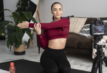 Bir kişi ev ortamında yoga matı üzerinde diz çökmüş pozisyonda, kolunu göğsünün önünde esnetiyor. Önünde tripod üzerinde bir kamera bulunuyor; bu da kişinin egzersiz kaydı veya çevrim içi içerik hazırladığını gösteriyor. Matın üzerinde bir su şişesi var. Arka planda koltuk, bitki ve merdiven görülen sıcak bir salon ortamı yer alıyor.