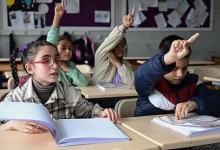 Görselin üst bölümünde, bir okul sınıfında sıralarda oturan ve derse katılmak için parmak kaldıran görme engelli öğrenciler yer alıyor. Ön planda sol tarafta, kırmızı çerçeveli gözlük takan, saçları örgülü ve ekose gömlekli bir kız çocuğu önündeki kabartma Braille alfabesiyle basılmış açık kitaba dokunarak dikkatle konuşuyor. Yanında oturan montlu erkek öğrenci sağ elini havaya kaldırıp diğer eliyle sıradaki kabartmalı çalışma kâğıdını takip ederken; arka sıralarda oturan diğer öğrenciler de derse katılmak amacıyla heyecanla parmak kaldırıyor.