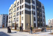 Modern bir sosyal konut yerleşimi görülüyor. Çok katlı, açık renk cepheli apartman bloklarının önünde geniş ve düzenli bir yaya alanı bulunuyor. Bu alanda tekerlekli sandalye kullanan bireyler, beyaz baston ve rehber köpek eşliğinde yürüyen bir kişi gibi farklı erişilebilirlik ihtiyaçlarına sahip insanlar rahatça hareket ediyor. Binaların girişleri düz zeminle bağlantılı, yollar geniş, peyzaj düzeni temiz ve engelsiz. Ortam, erişilebilirlik ilkelerine uygun tasarlanmış bir sosyal konut projesinin kapsayıcı yapısını yansıtıyor; herkesin bağımsız ve güvenli biçimde dolaşabildiği, eşit kullanım imkânı sunan bir yaşam alanı hissi veriyor.