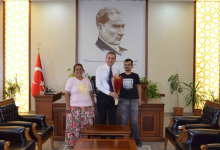 Görselin üst bölümünde, makam odasında gerçekleşen tebrik ziyaretinden neşeli bir an yer alıyor. Ortada beyaz gömlekli ve kravatlı rektör/yetkili, elinde kırmızı-mor renkli kuru çiçek buketini tutarak dururken; solunda açık pembe tişörtlü ve çiçek desenli etek giymiş tebessüm eden anne, sağında ise siyah baskılı tişört ve kot pantolon giymiş görme engelli üniversite öğrencisi Özcan Şaylı yer alıyor. Üçlü, arkalarında ahşap çalışma masası, büyük boy Mustafa Kemal Atatürk portresi ve Türk bayrağı ile yanlardaki deri koltukların bulunduğu resmi makam odasında yan yana kameraya gülümsüyor.