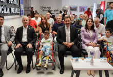 Görselin üst bölümünde, modern bir konferans veya etkinlik salonunda yan yana dizilmiş sandalyelerde oturan protokol üyeleri, davetliler ve özel gereksinimli çocuklar yer alıyor. Ön sırada, bacaklarında yürüme cihazı (ortez) bulunan ve tekerlekli sandalyesinde oturan küçük bir erkek çocuğu; solunda ve sağında takım elbiseli erkek yöneticiler ile açık lila ceketli kadın yetkili oturarak kameraya bakıyor. En sağda ise yine kırmızı tekerlekli sandalyede oturan gözlüklü başka bir çocuk görülüyor. Arkalarındaki sıralarda aileler ve diğer katılımcılar yer alırken, arka duvarda "İyiliğe değil, İyileştirmeye geldik Hatay 2024" yazısı ve sol tarafta duvarda asılı merkezin tabelası seçiliyor.
