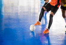 Görselde, kapalı bir sahada turuncu formalı bir futbolcunun top sürerken, rakibinin ise onu takip ettiği bir futsal maçı görülüyor.
