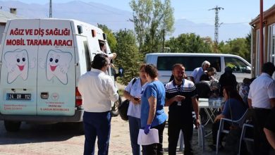 Açık havada, dağların göründüğü bir alanda mobil bir diş sağlığı tarama aracı bulunuyor. Aracın üzerinde “Ağız ve Diş Sağlığı Mobil Tarama Aracı” ve “T.C. Sağlık Bakanlığı Erzincan İl Sağlık Müdürlüğü” yazıları yer alıyor. Yanında gülümseyen iki çizgi film tarzı diş karakteri görülüyor. Aracın önünde ve yanında birkaç kişi ayakta sohbet ediyor; gölgeli bir bölümde ise masalar kurulmuş, bazı kişiler oturarak işlem veya bilgilendirme alıyor gibi görünüyor. Ortam bir halk sağlığı etkinliğini, yerel topluma yönelik ücretsiz bir tarama hizmetini yansıtıyor. Genel atmosfer sıcak, toplumsal faydaya yönelik ve açık hava etkinliği havasında.