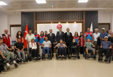 Instagram İçin Alternatif Metin (Akülü Sandalye Desteği Haberi): Görselin üst bölümünde, resmi bir kurumun toplantı veya kabul salonunda düzenlenen teslim töreninde bir araya gelen kalabalık bir grup yer alıyor. Ön sırada siyah renkli yeni akülü tekerlekli sandalyelerine oturmuş farklı yaş gruplarından kadın ve erkek engelli bireyler yan yana sıralanıyor; arka sırada ise takım elbiseli mülki idare amirleri, protokol üyeleri ve kırmızı-mavi yelekleriyle Aile ve Sosyal Hizmetler Bakanlığı görevlileri ayakta durarak kameraya toplu hatıra pozu veriyor. Arka planda Türk bayrağı, bakanlık logosu ve ahşap kaplamalı modern salon duvarı görülüyor.