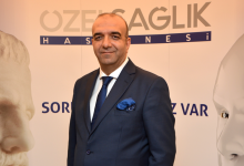 Görselde takım elbiseli Op. Dr. Kenan Kalı, üzerinde “Özel Sağlık Hastanesi” yazılı bir tanıtım panosunun önünde duruyor. Arka planda iki büyük heykel yüzü yer alıyor. Ortam resmi ve kurumsal bir sunum alanı izlenimi veriyor.