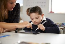 Sınıf ortamında, bir yetişkin ile bir çocuk yan yana oturuyor. Yetişkin tablet ekranını işaret ederken çocuk kalemle ekrana odaklanmış durumda. Arka planda sınıf eşyaları flu şekilde görülüyor.