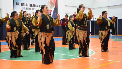Fotoğrafta, bir spor salonunda sahne alan özel gereksinimli bireylerden oluşan geleneksel halk dansı ekibi görülüyor. Dansçılar, altın sarısı gömlekler, siyah yelekler ve siyah pantolonların üzerinde alev formunda altın detaylar bulunan uyumlu kostümler giymiş durumda. Spor salonunun zemini turuncu ve yeşil renkli bir saha çizimiyle kaplı. Arka planda büyük bir afiş yer alıyor. Afişte bir erkek portresi ve “DR. HALİL İBRAHİM ASLAN – BELEDİYE BAŞKANI” yazısı bulunuyor. Bu detay, etkinliğin bir belediye tarafından düzenlenen kültürel veya toplumsal bir organizasyon olduğunu düşündürüyor. Dansçılar senkronize bir şekilde poz verirken, ortamın genel atmosferi coşkulu ve geleneksel bir gösteri havası taşıyor.
